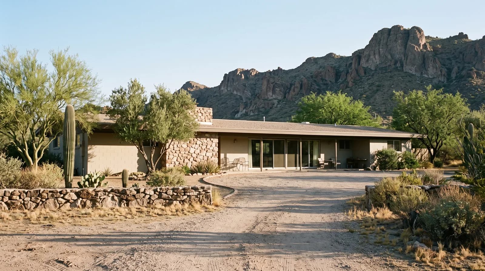 A low 1960s desert ranch house set back on a large lot, with a gravel motor court and a rocky ridge behind